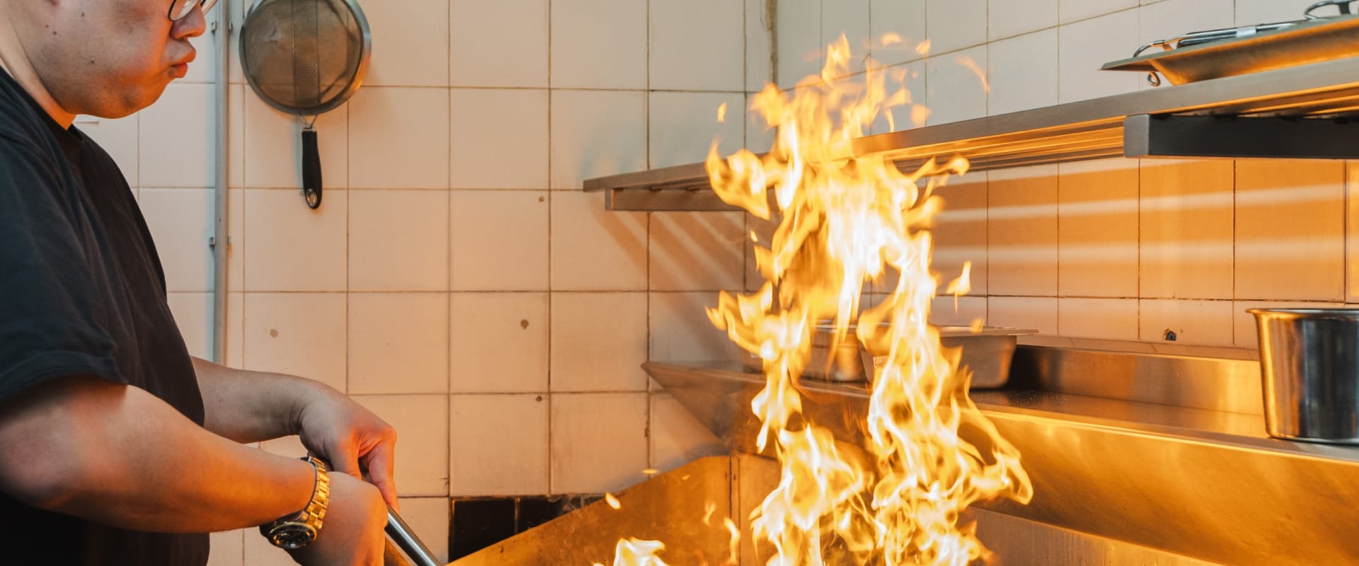 Chef stir-frying with a wok in a stainless steel kitchen, creating a tall blaze of fire.