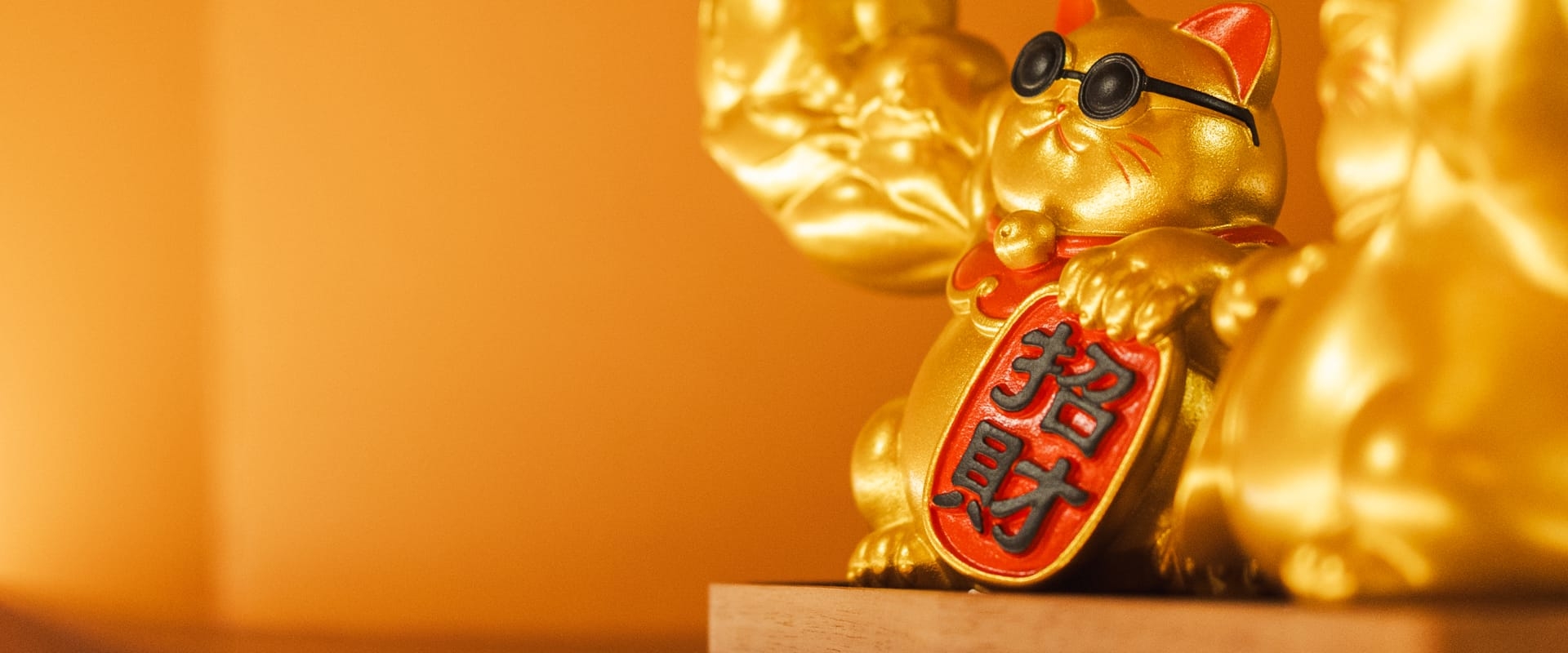 Golden Maneki-neko cat figurine wearing small black glasses on an orange background.