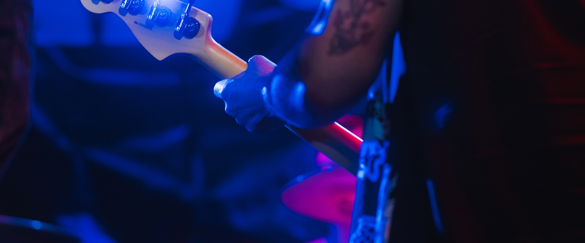 Musician's hand playing a bass guitar neck on a dark stage with intense blue and pink lighting.